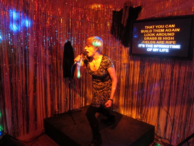 Women with short blonde hair singing karaoke at Apexart's SCARYOKE art exhibition