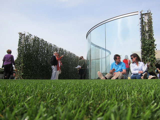 An image of two people sitting against Dan Graham's two-way mirror installation on the Met's rooftop garden