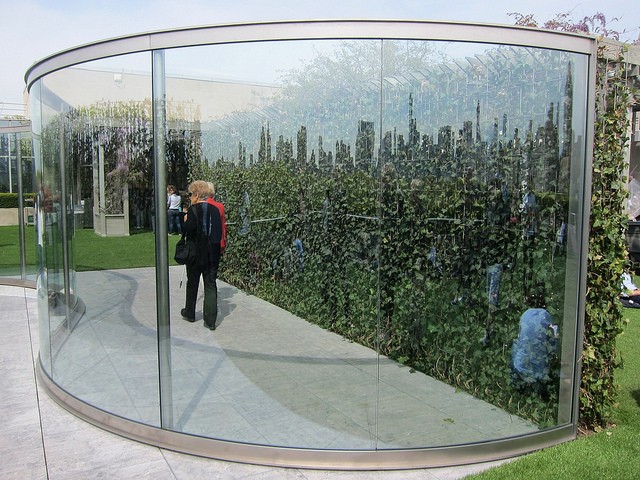 A women walks within the Met's rooftop installation with a wall of ivy and a two way mirror wall