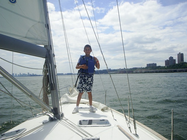 A young boy with a blue life jacket on stands at the tip of the sail boat on his first sailing lesson in NYC.