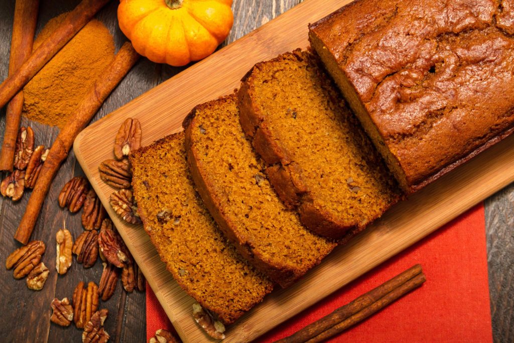 Pumpkin bread sliced on a table with pumpkin, cinnamon and nuts in the background.